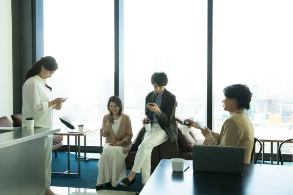 オフィスの休憩スペースで、複数人がスマートフォンやパソコンを使って作業や情報確認をしている様子